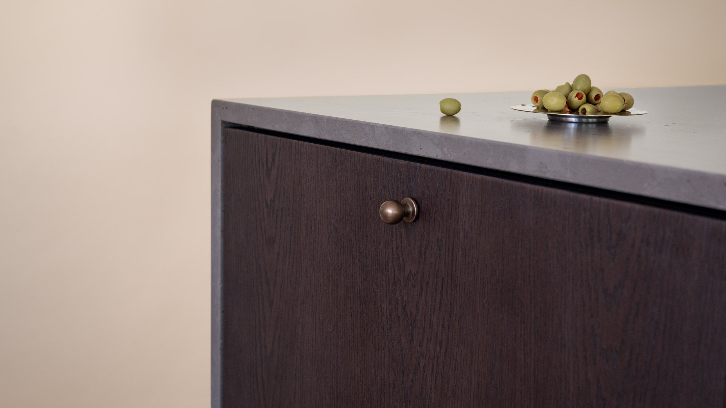A close-up of a cabinet featuring Fronteriors Charred Oak doors with a matte gray countertop and a round brass Hepburn Hardware knob, set against a warm beige wall with olives on a small plate on top.